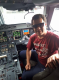 Descrição da imagem: Homem de pele clara e cabelo castanho escuro, usando óculos escuros, sentado sorrindo no banco da direita (lado do copiloto) da cabine de comando de um Embraer 195 E1 da companhia aérea Azul. Ele veste camiseta vermelha com a frase “APRENDI A VER COM O CORAÇÃO” em branco e calça azul escura. A cabine está bem iluminada pela luz natural que entra pela janela lateral, destacando o rosto dele e os painéis e telas digitais do cockpit ao fundo. Fim da descrição.